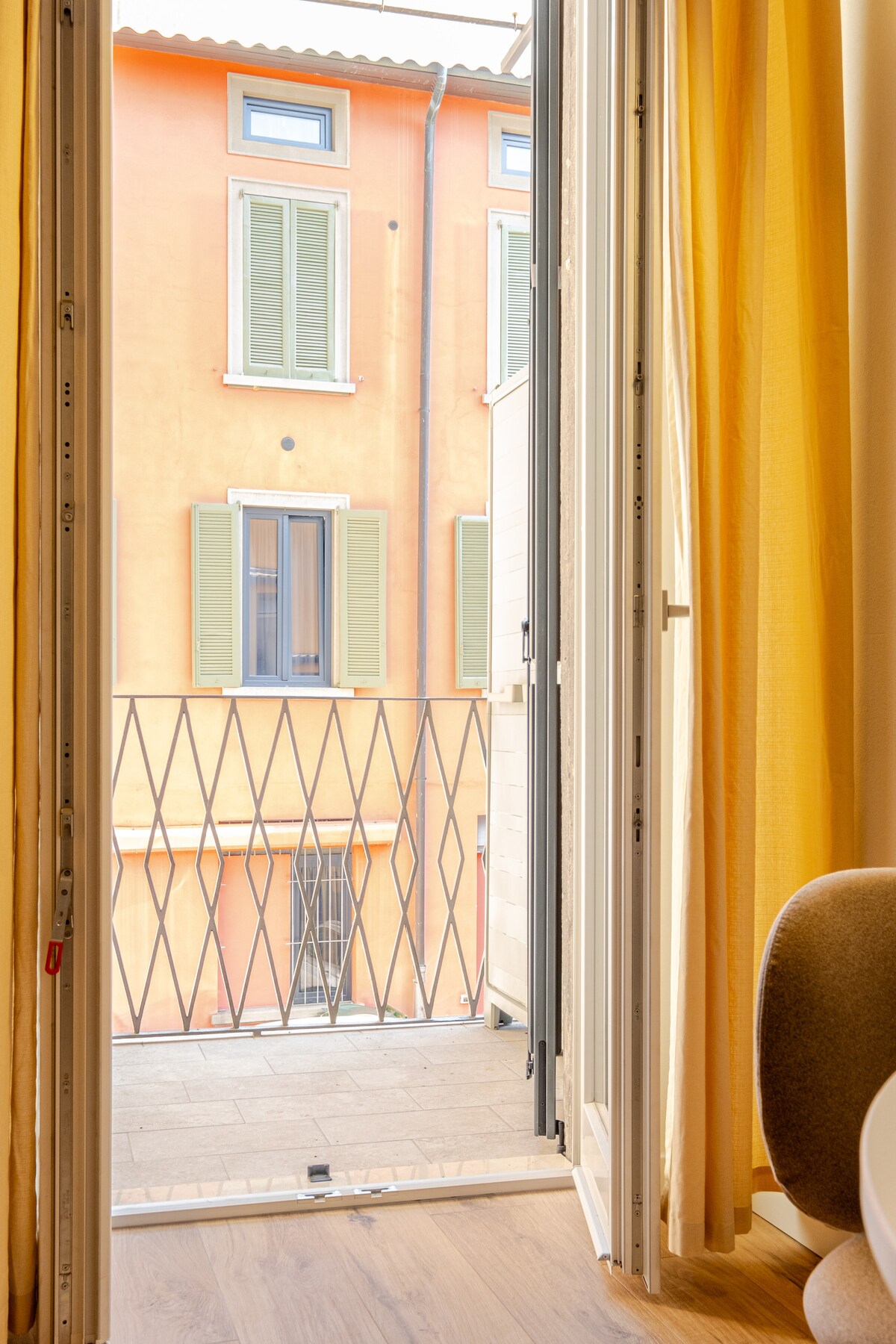 A French door opens to a small balcony, offering a view of a vibrant orange building with green shutters. Natural light filters through yellow curtains, enhancing the warm ambiance of the space.
