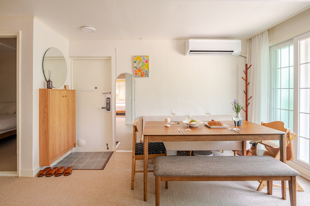 A dining area features a wooden table set for a meal, complemented by a minimalist bench and chairs. Light streams in through large windows, illuminating the neutral-toned walls and soft furnishings. A small mirror and artwork add subtle decorative elements to the space.
