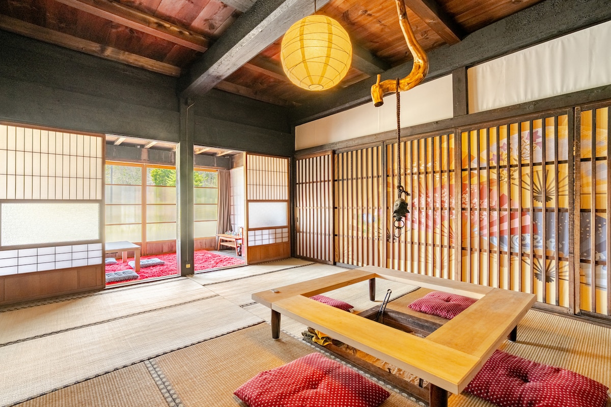 A traditional Japanese room features tatami flooring, wooden beams, and shoji screens that allow natural light to filter through. A low wooden table sits in the center, accompanied by red cushions. The space maintains a serene ambiance with a lantern hanging from the ceiling.