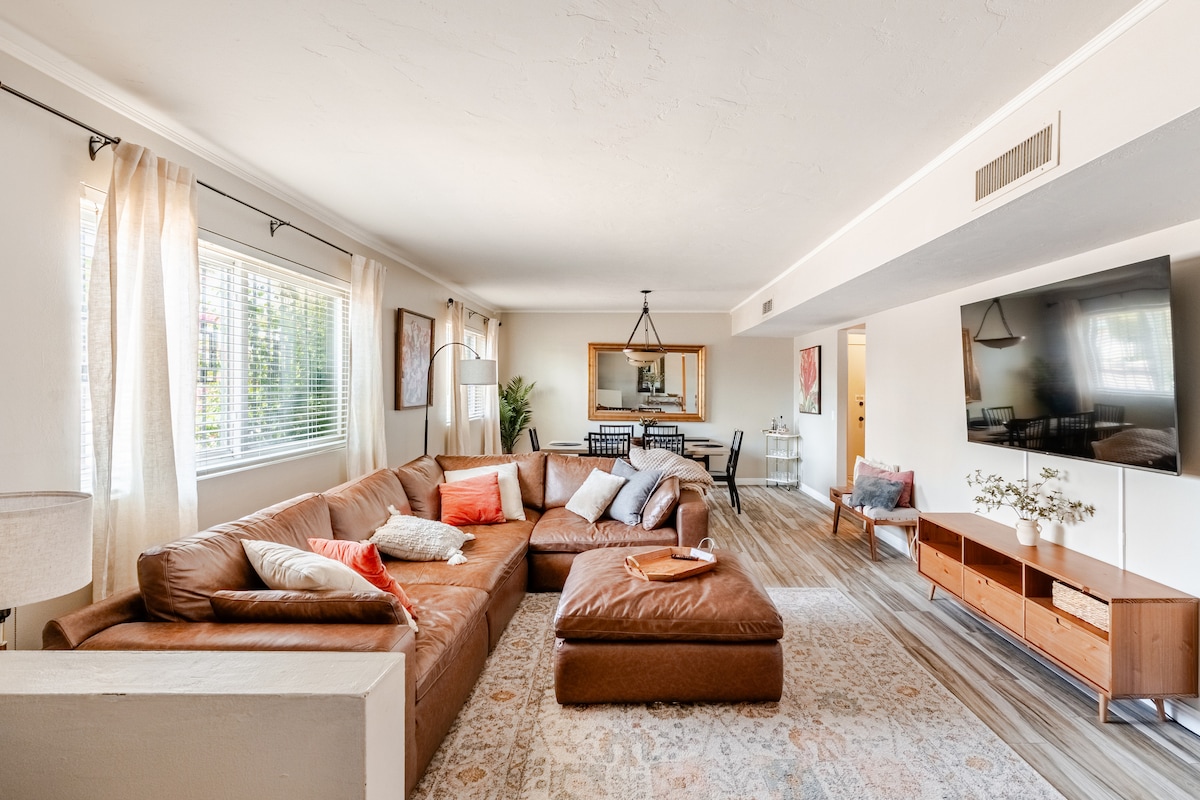 A spacious living room is featured with a large brown sectional sofa adorned with pillows in various colors. A flat-screen TV is mounted on the wall across from the seating area. Nearby, a dining table is set for six, and natural light filters through the window.