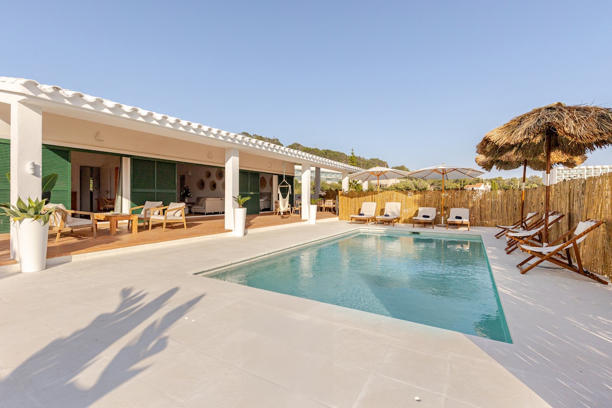 A private pool is surrounded by a spacious sun terrace featuring multiple loungers and a shaded seating area. The villa's modern architecture is complemented by natural elements, while lush greenery frames the outdoor space, creating a serene environment for relaxation.