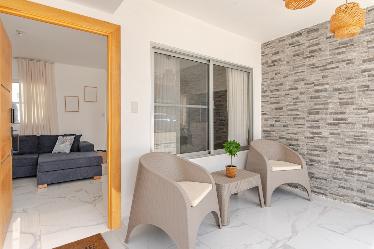 A welcoming entryway is presented with two light-colored chairs and a small table, complemented by a decorative plant. The textured stone wall behind adds visual interest, while natural light filters through large windows, providing a bright and airy atmosphere.