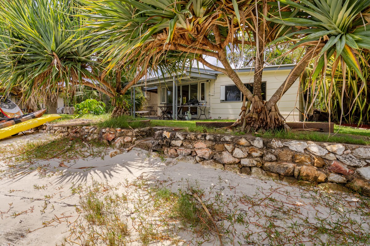 A cabin is nestled amidst lush palm trees, with a backdrop of a sandy shoreline leading to the water. The outdoor area features a stone wall and a patio with seating, inviting guests to enjoy the serene surroundings. A kayak is positioned on the beach.