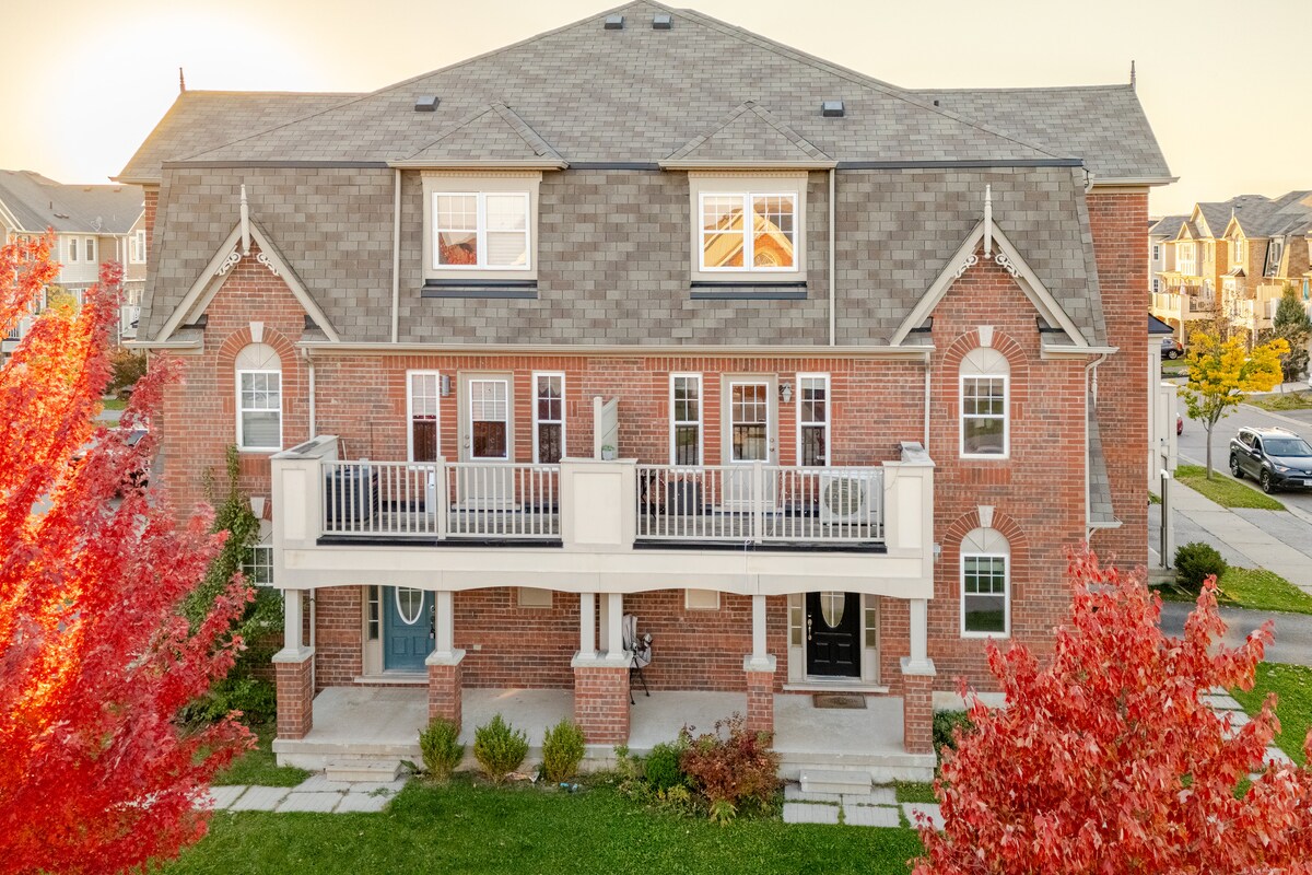 A charming two-story townhouse is presented with red-brick exterior, flanked by vibrant autumn trees. A spacious balcony is situated above the entrance, offering outdoor seating. The well-maintained front garden and walkway enhance the inviting atmosphere of the property.