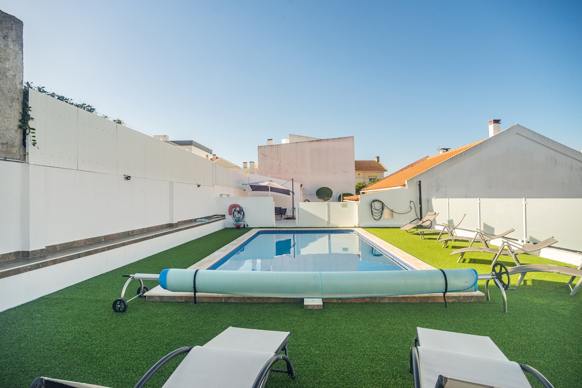 A clear view of a swimming pool surrounded by well-maintained green grass. Sun loungers are arranged neatly on one side of the pool, while a white cover is rolled along the edge of the pool. The area is enclosed by walls, offering a private atmosphere.
