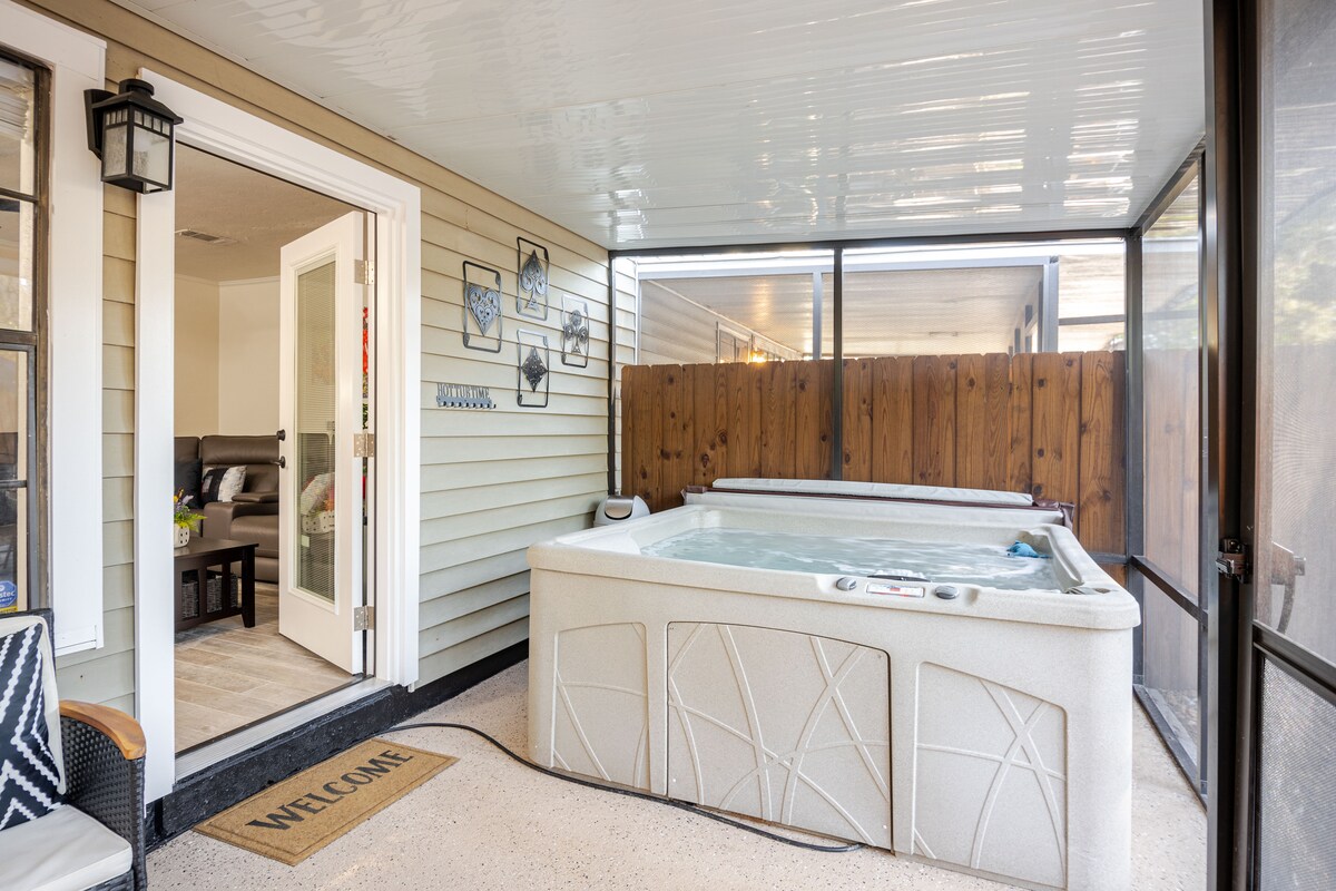 A screened-in back porch features a hot tub for four, complemented by light-toned walls and a welcoming entry mat. The space offers a view of the enclosed yard beyond the wooden fencing, creating a private and relaxing atmosphere.