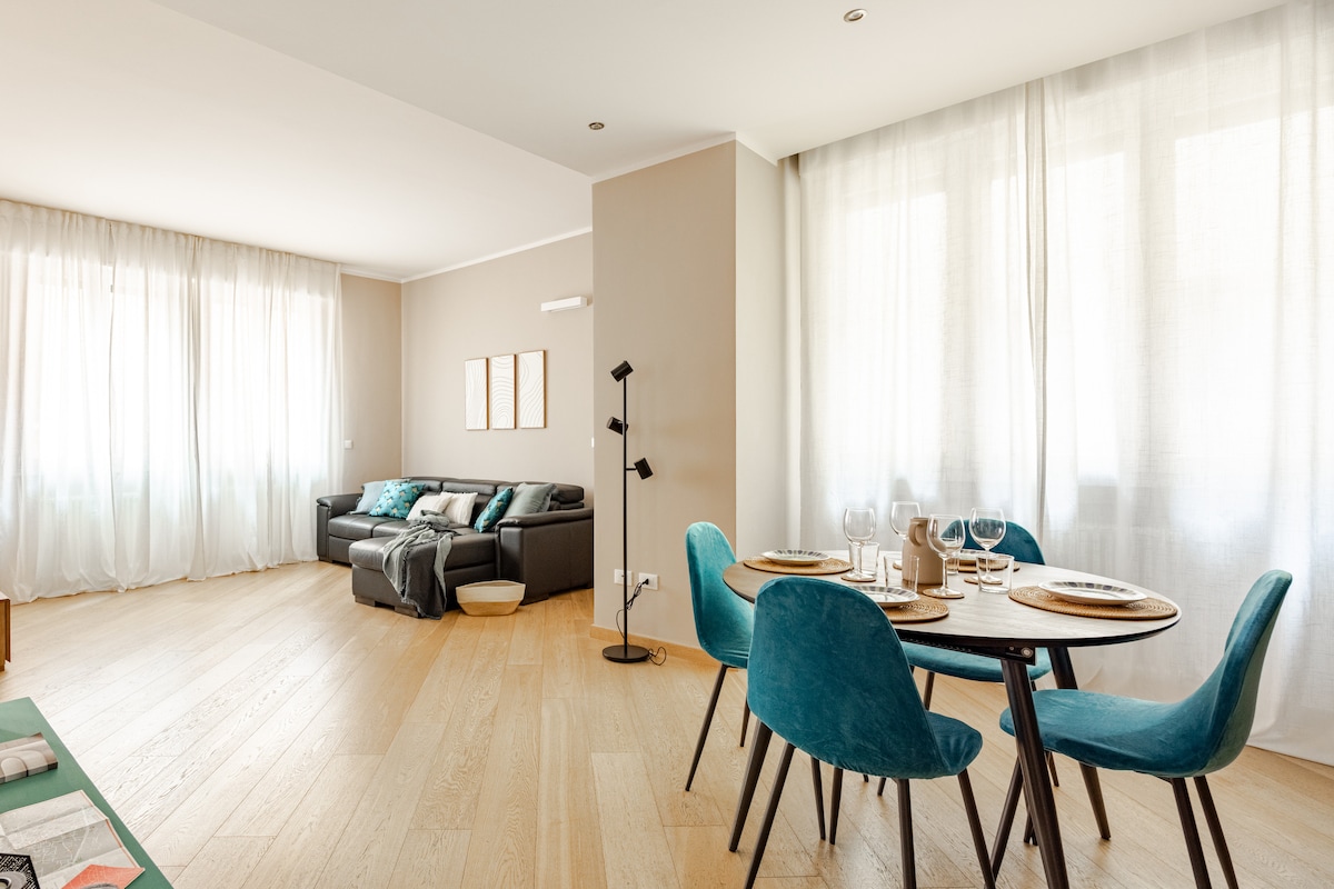 A spacious living area is highlighted by large windows with sheer curtains, allowing natural light to fill the room. A comfortable sofa is arranged with decorative cushions, while a modern dining table with teal chairs is set for a meal, enhancing the inviting atmosphere.