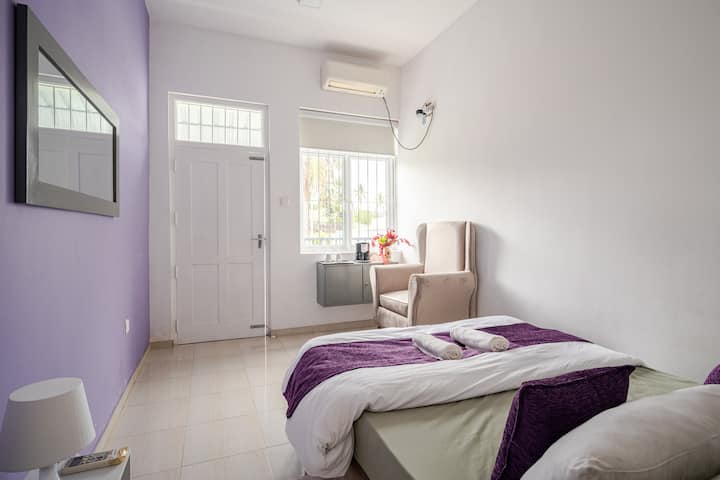 Violet Twilight-Cosy bedroom featuring a comfortable grey bed with elegant purple accents with A/C for a relaxing stay. A private balcony passage leads to the rear side of the property, overlooking a quiet street with a refreshing green view.