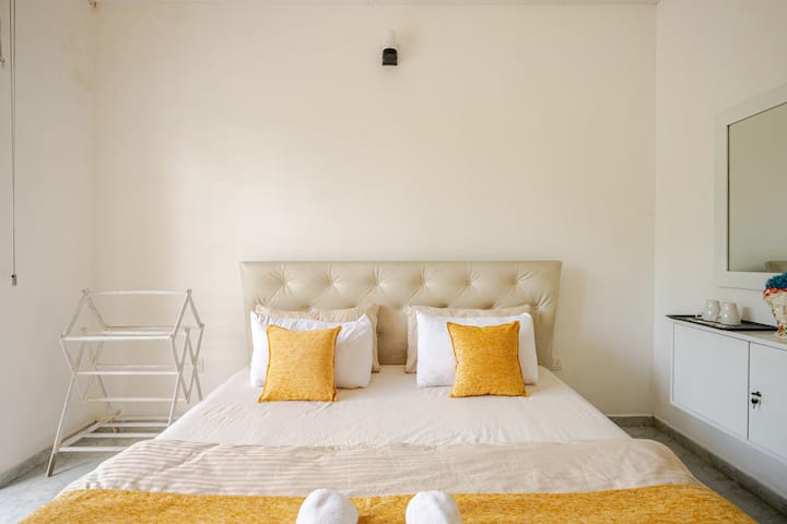 A spacious first-floor room with a large bed, posture-support mattress ideal for elderly guests, air conditioning, and French windows opening to the entrance garden and blue sky.