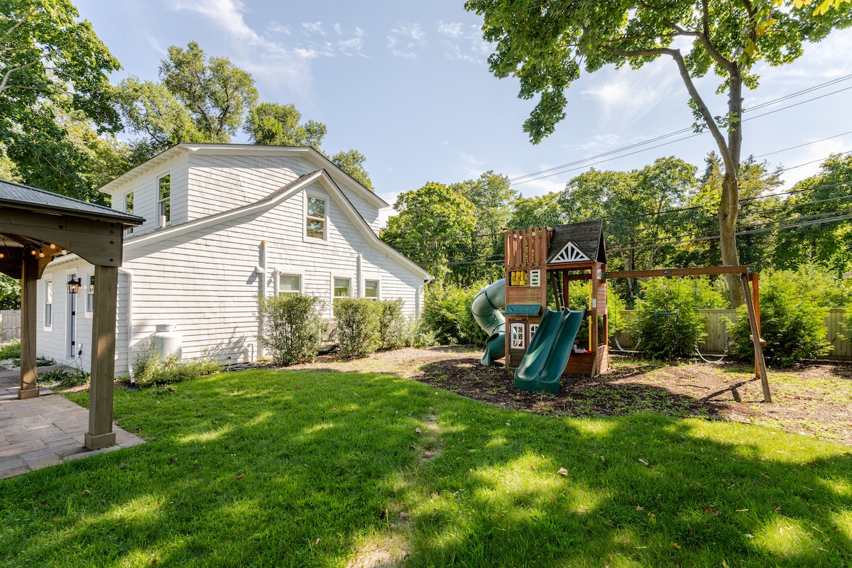 A spacious grassy area is highlighted by a playground structure that includes a slide, climbing features, and a small fort. The house with white siding is visible in the background, surrounded by lush greenery, creating an inviting outdoor play space.