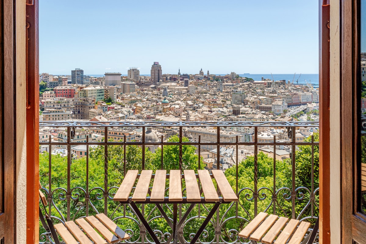 A small wooden table with two matching chairs is positioned on a balcony, offering expansive views of Genoa's cityscape. The scene captures the rooftops and distant coastline under a clear blue sky, framed by ornate balcony railing and open double doors.
