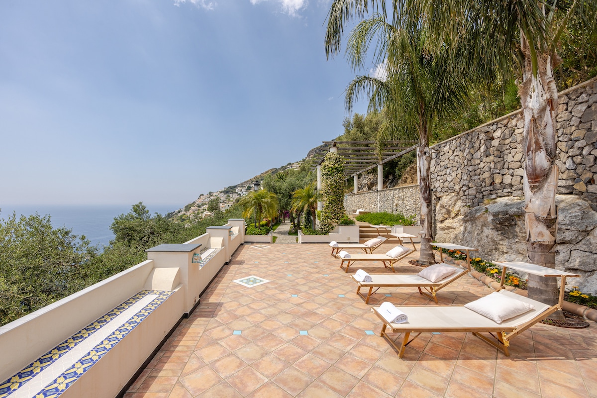 A spacious terrace with sun loungers is highlighted by warm, terracotta tiles. Lush greenery and garden beds surround the area, providing a sense of tranquility. A stone wall creates a natural backdrop, with ocean views visible in the distance under a bright sky.