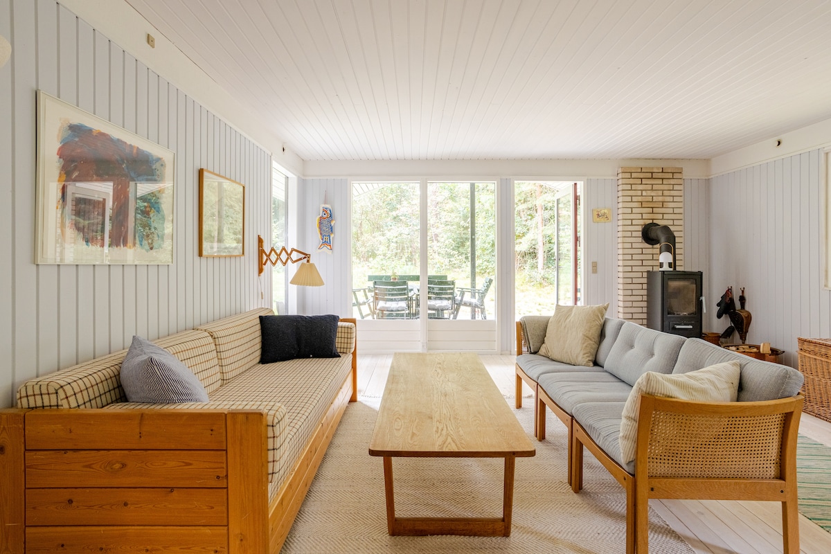 A bright living area is depicted with light-colored walls and a textured rug underfoot. Two comfortable sofas are arranged around a wooden coffee table. Large glass doors lead to an outdoor seating area, allowing natural light to fill the space.