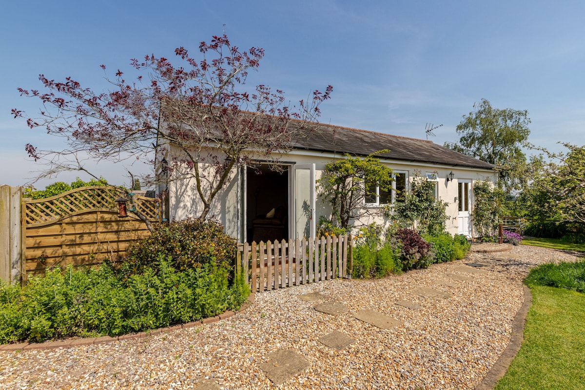 The exterior of the studio apartment is shown, featuring a gravel pathway leading to a white building surrounded by greenery. A wooden fence with a gate provides access, complemented by flowering plants and trees that enhance the welcoming appearance.