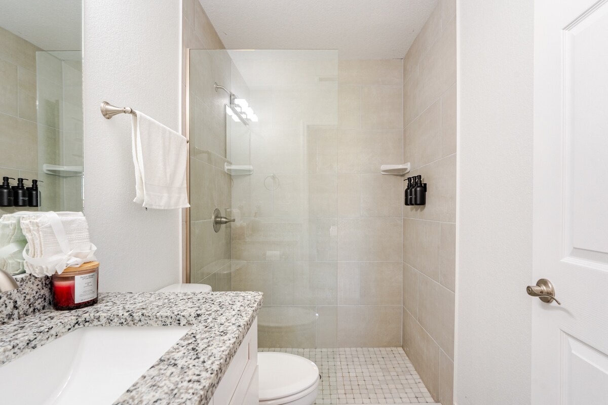 A modern bathroom is displayed with a glass-enclosed shower. Natural light enters through a frosted glass panel. A spacious vanity with a granite countertop is visible, complemented by neatly arranged toiletries and a white towel hanging nearby. The overall design emphasizes a clean and minimalist aesthetic.
