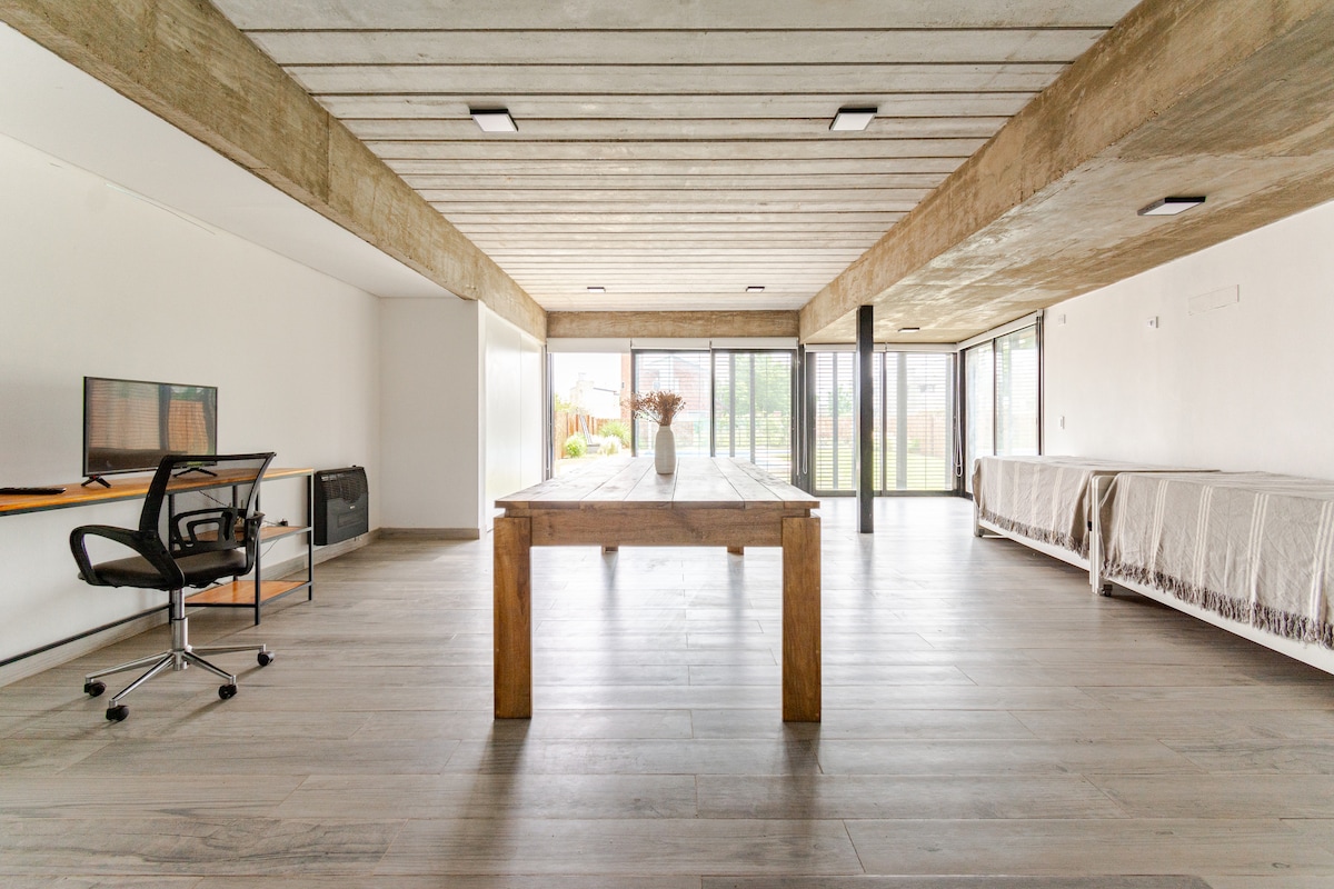 A spacious living area is characterized by concrete ceilings and large glass doors that bring in natural light. A wooden table stands prominently in the center, surrounded by a neutral color palette. Comfortable chairs are positioned nearby, with views to the outdoor patio.