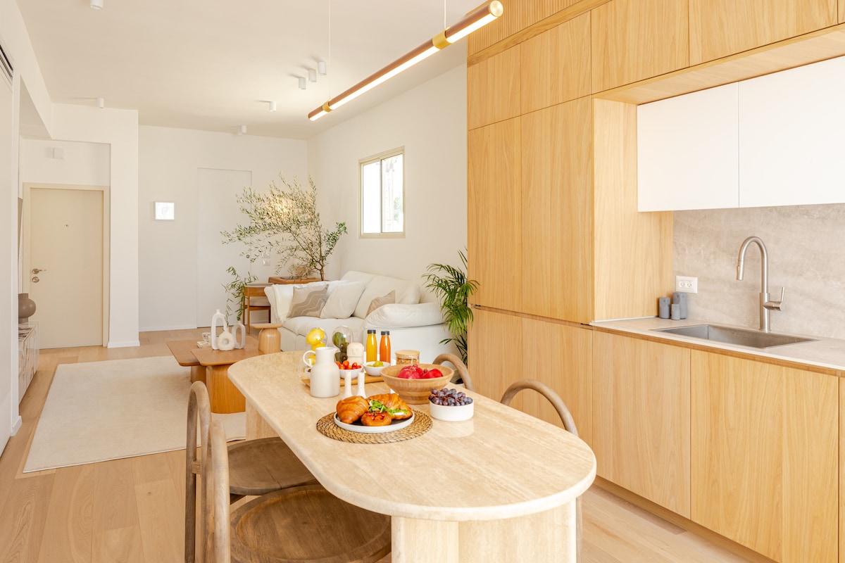 A dining area is seen with a light wooden table set for breakfast, featuring pastries and drinks. In the background, a spacious living area includes a comfortable, light-colored sofa with greenery and a soft rug. Natural light brightens the space through large windows.