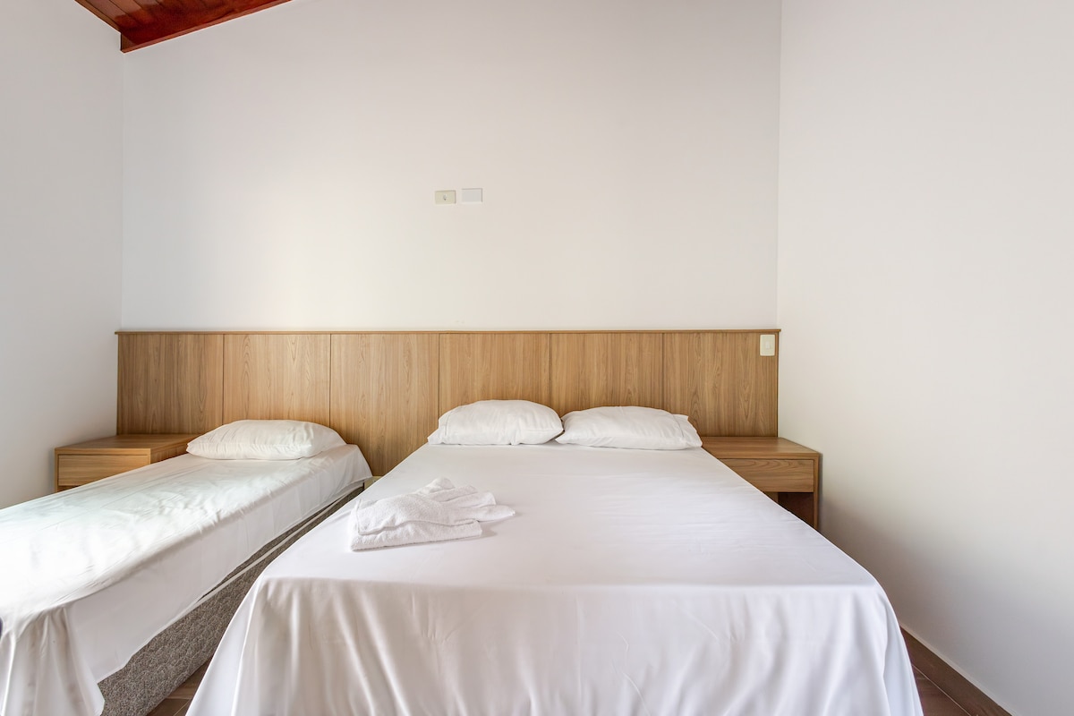 The room features a spacious bed accompanied by a smaller bed. Wooden paneling creates a simple and clean backdrop, while natural light illuminates the area. Two bedside tables are positioned on either side of the larger bed.