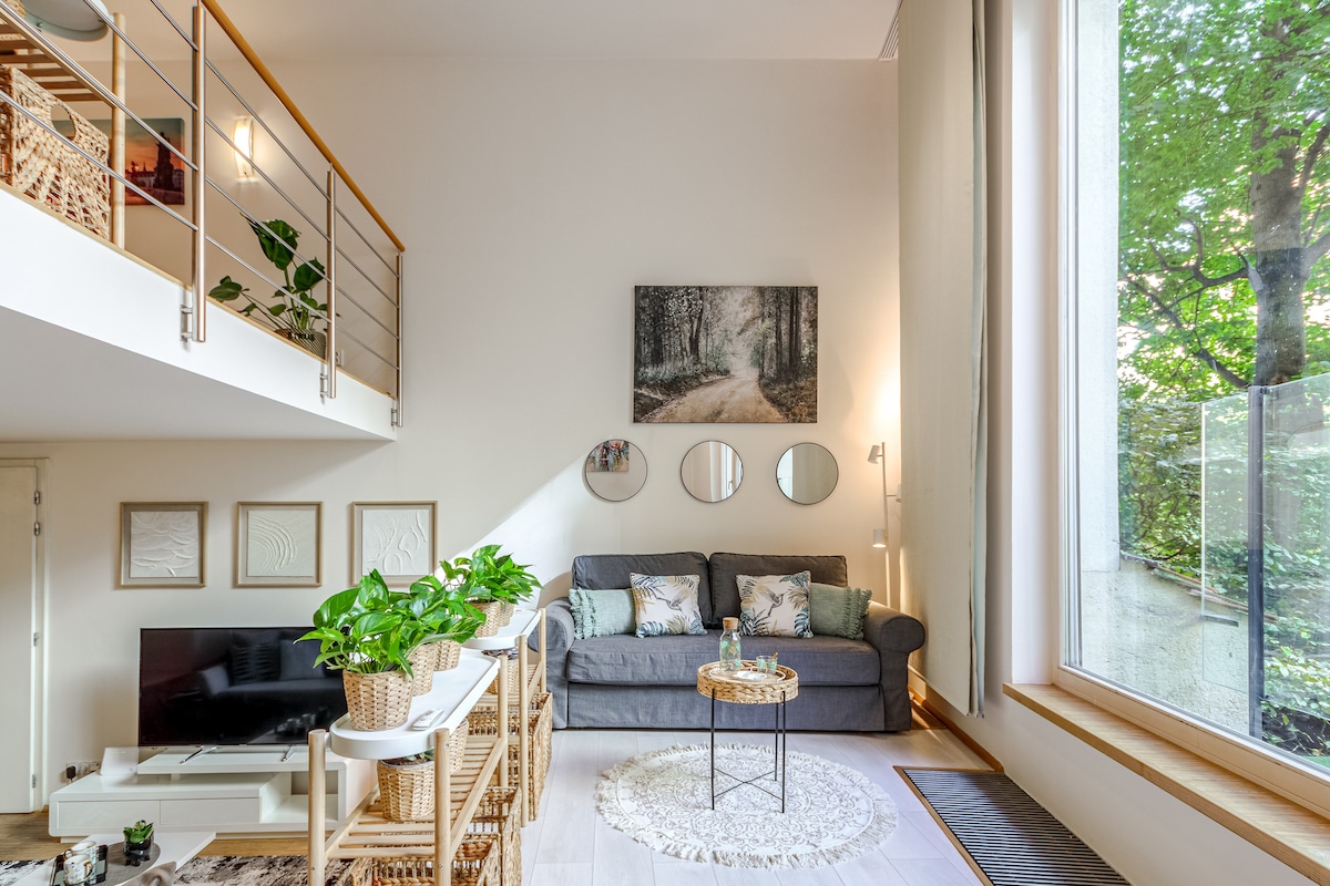The living area features a comfortable gray sofa and a round coffee table, offering a cozy setting. Large windows allow natural light to brighten the space and provide views of greenery outside. Decorative elements include wall art and indoor plants, enhancing the inviting atmosphere.