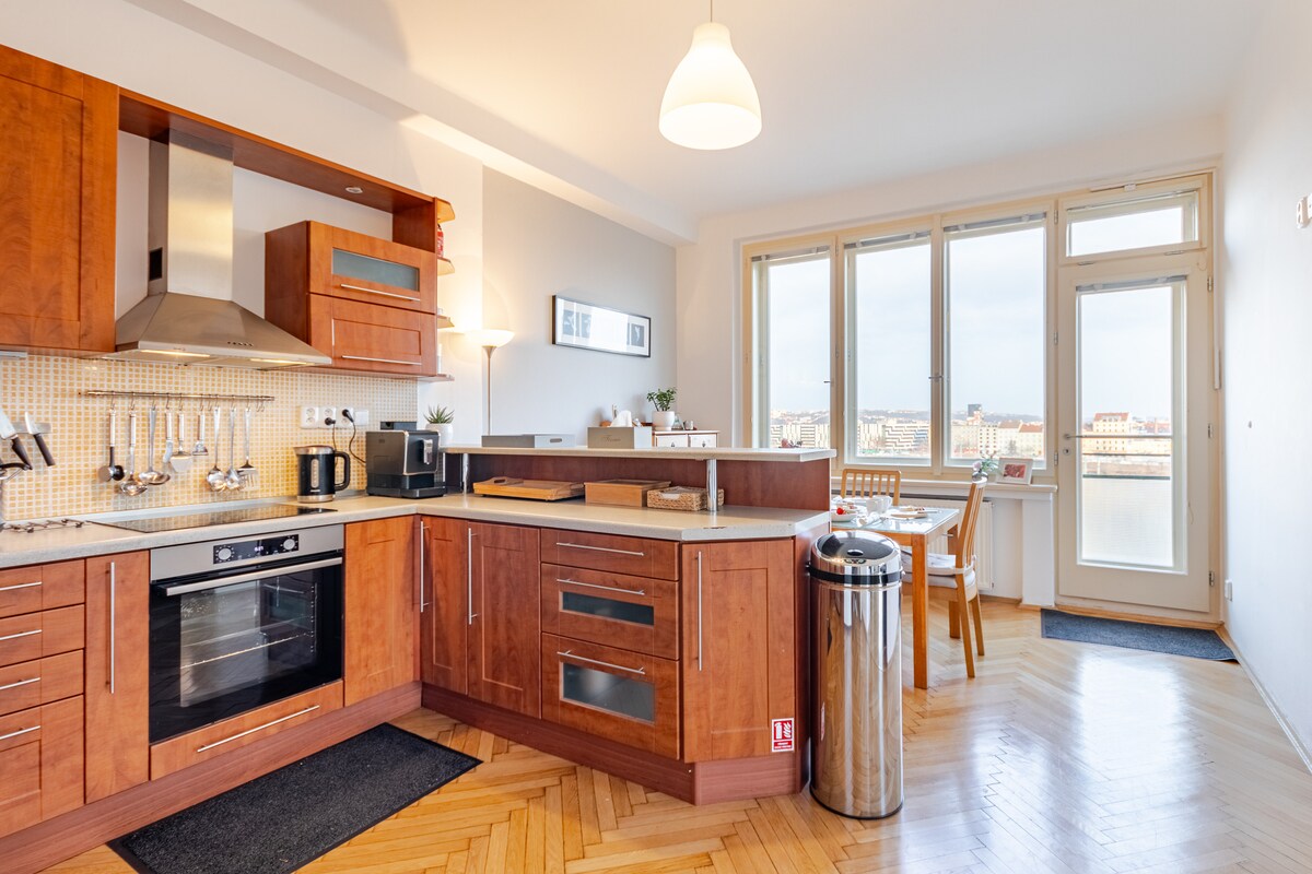 The kitchen features modern wooden cabinetry and stainless steel appliances, including an oven and microwave. A dining area is nearby, with a table and chairs positioned by large windows that provide ample natural light and a view of the surrounding area.