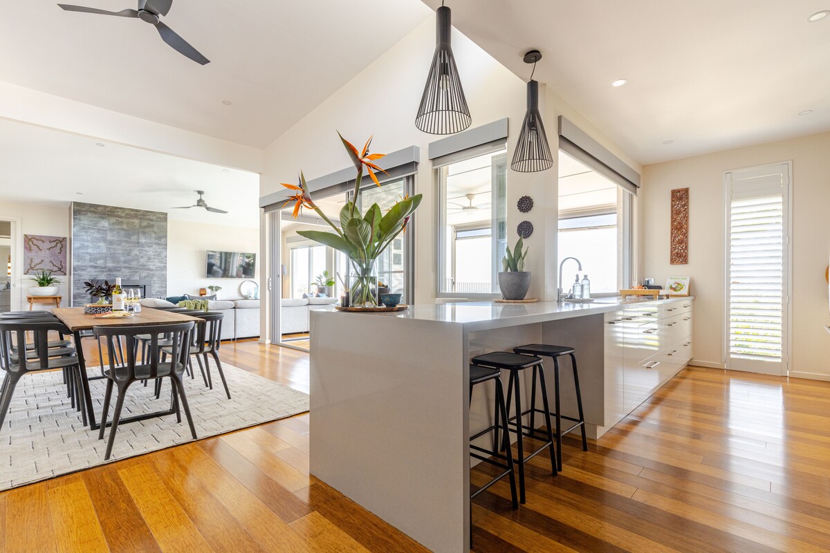 A spacious kitchen features a large island with seating for three and is adorned with tropical plants. Natural light streams in through expansive glass doors, while stylish pendant lights hang above. The seamless flow into the dining area showcases a modern dining table set against warm wooden flooring.