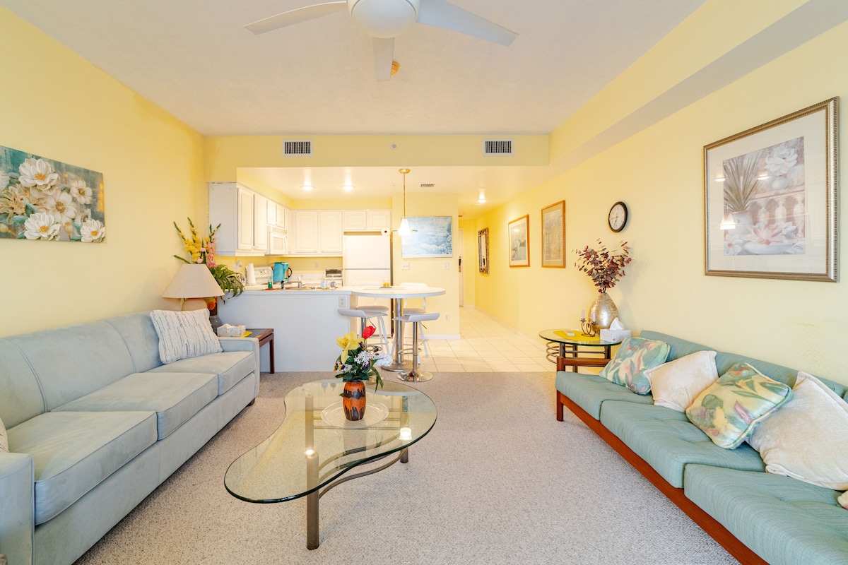 A spacious living area is presented with soft yellow walls and plush seating arrangements. A glass coffee table sits at the center, adorned with a small flower arrangement. The open kitchen area is visible in the background, featuring bright cabinetry and a dining table.