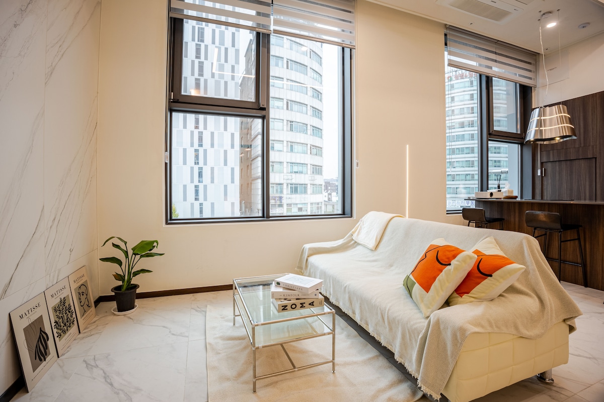 A bright living area is depicted, featuring large windows that allow natural light to flood in. The space includes a white sofa adorned with colorful cushions and a minimalist coffee table. A small plant adds a touch of greenery, while modern artwork is displayed on the wall.