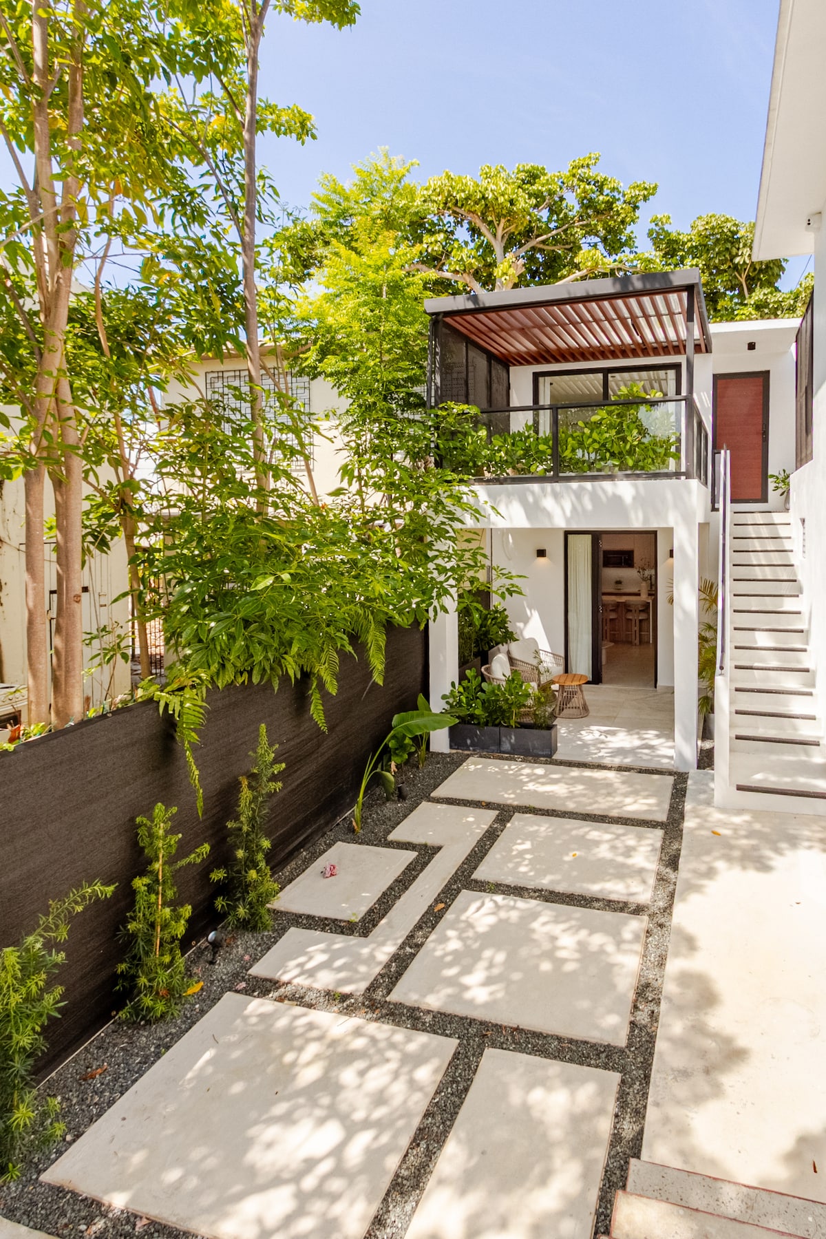 A welcoming entrance showcases a landscaped patio with stone pathways leading to the studio. Lush greenery surrounds the area, providing a sense of privacy. A staircase on the right leads to the upper level, while an inviting doorway opens into the interior of the home.