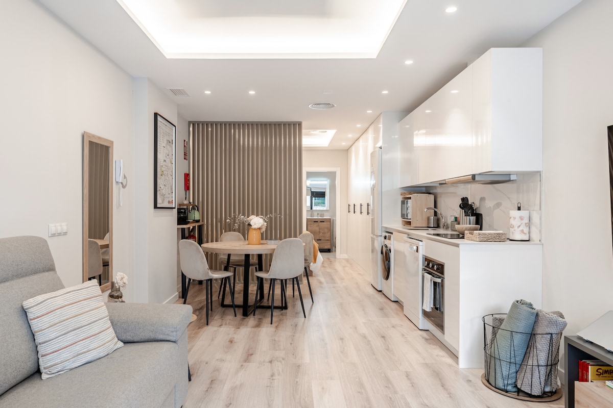 The living area is highlighted by a light grey sofa and a dining table with four chairs. A sleek kitchen with a modern design and integrated appliances is visible. Soft lighting enhances the open layout, creating a warm and spacious environment.