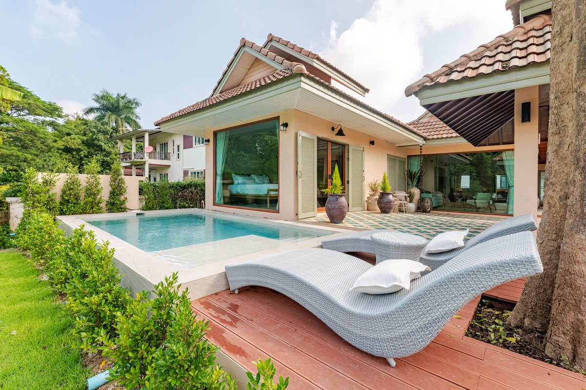 A modern villa is showcased with a private pool surrounded by greenery. Lounge chairs are positioned beside the pool, providing a relaxing space. Large windows and doors allow ample natural light, connecting the indoor and outdoor areas. The peaceful setting invites guests to unwind.