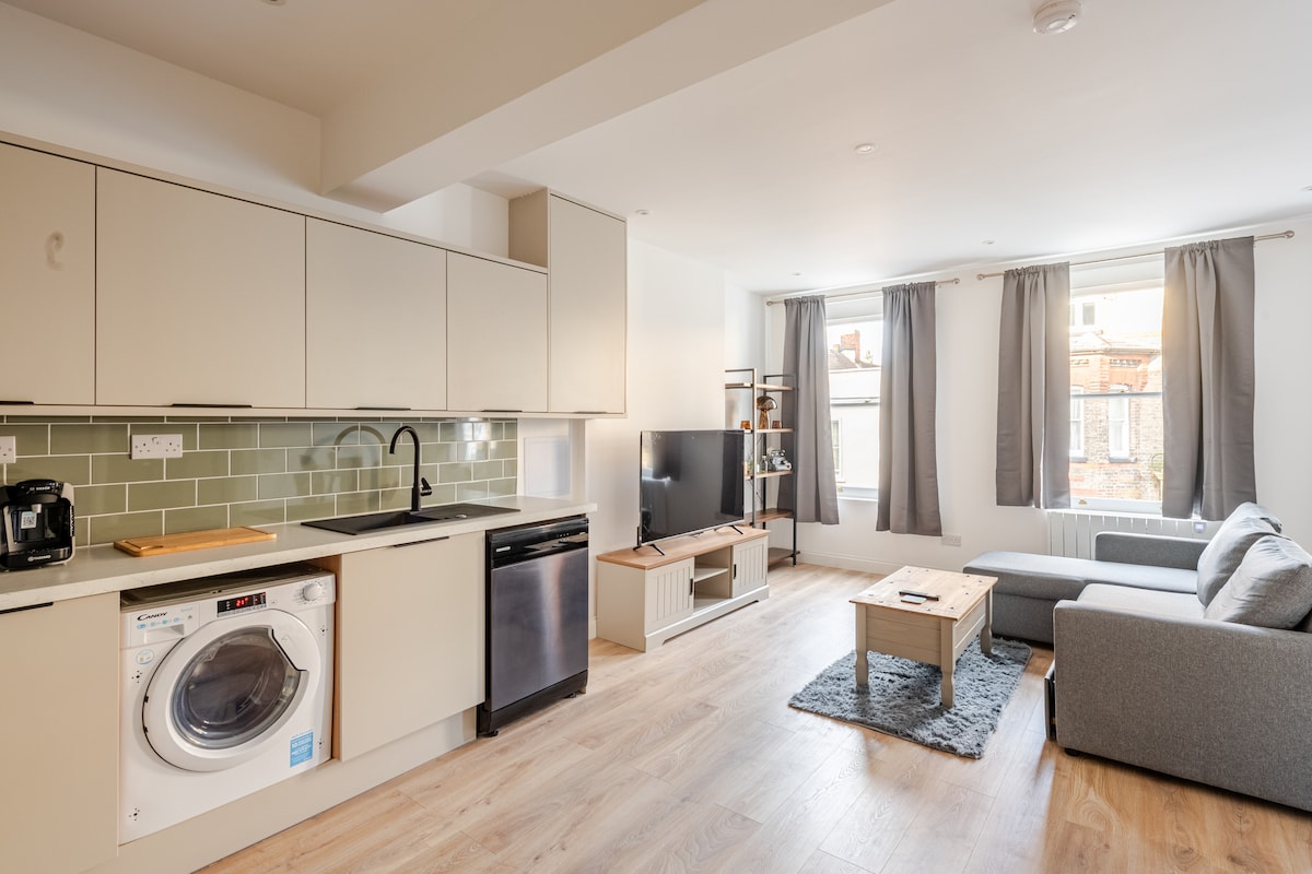 A modern kitchen is integrated into an open living space, featuring light-colored cabinetry, a washing machine, and a black dishwasher. A cozy seating area includes a gray sectional sofa and a wooden coffee table, with natural light filtering through two large windows adorned with gray curtains.