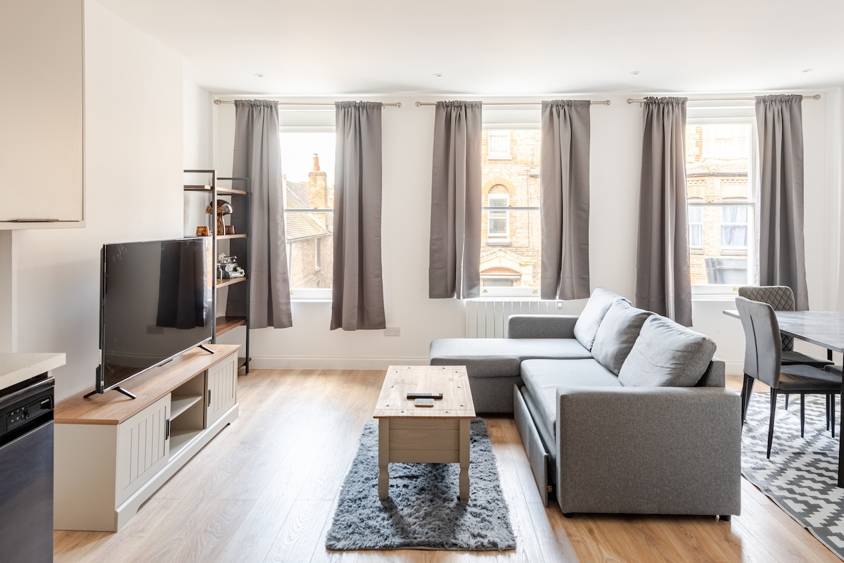 A modern living area features a comfortable gray sectional sofa and a wooden coffee table atop a soft area rug. Large windows with gray curtains allow natural light to fill the space, while a TV is mounted on a contemporary media console. A dining table is visible in the background.