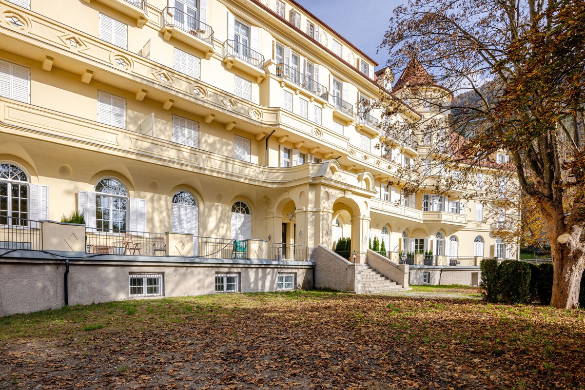 A serene building's exterior is displayed, characterized by its classic architecture. The façade features multiple arched windows and balconies, surrounded by a well-maintained lawn scattered with autumn leaves. Trees provide a natural frame, enhancing the peaceful outdoor environment.
