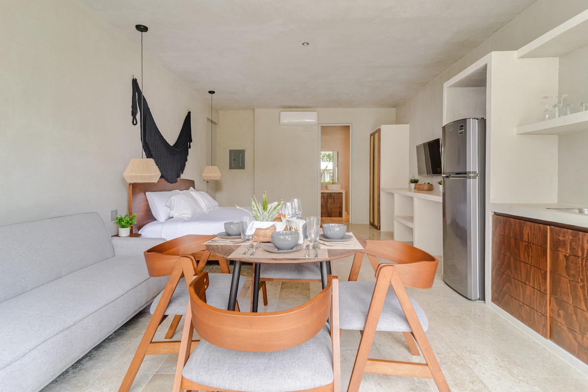 A modern living space features a comfortable gray sofa and a dining table surrounded by wooden chairs. Light wood cabinetry and stainless-steel appliances complement the serene atmosphere. Natural light fills the area, highlighting minimalistic decor and a spacious layout.
