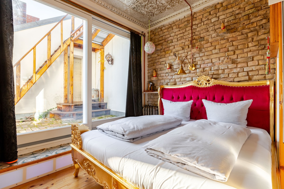 A spacious bedroom features a grand bed with a vibrant pink headboard and classic decor. Large windows allow natural light to illuminate the room, offering a view of an outdoor area with stone paving. Soft bedding is arranged neatly atop the bed.