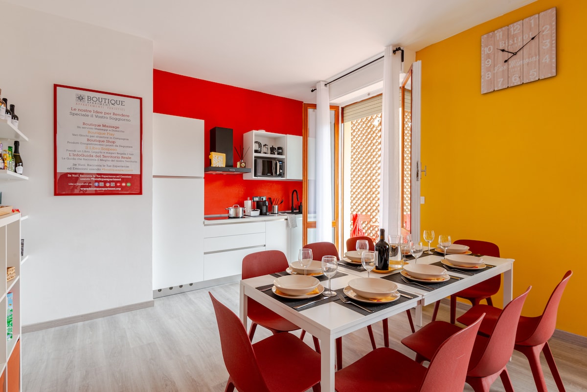 A modern living space features a dining table set for six, surrounded by red chairs. The kitchen area, equipped with essential appliances, includes contrasting walls in white and vibrant colors. Natural light filters through sliding doors, leading to an outdoor terrace.