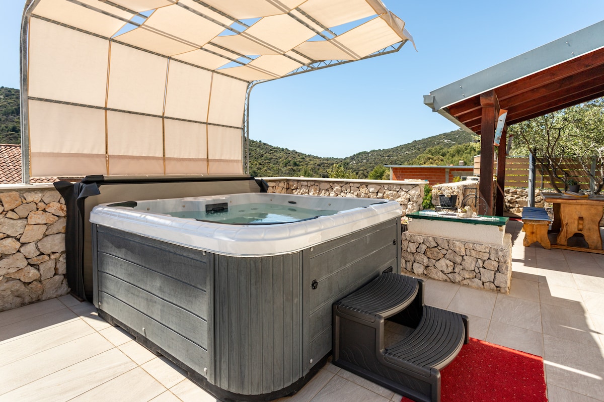 A spacious jacuzzi is set on a tiled terrace, offering a relaxing spot under a retractable canopy. Steps lead up to the hot tub, surrounded by a natural stone wall and views of greenery in the background.