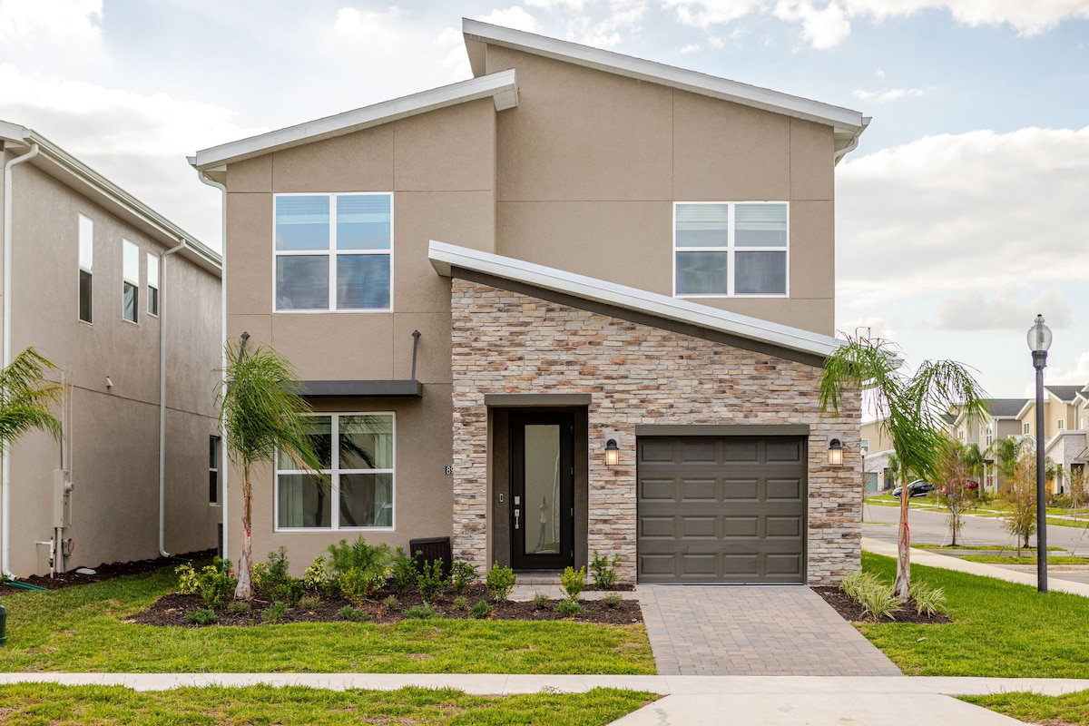 A modern two-story villa features a combination of light stucco and stone accents. Large windows allow natural light and enhance the sleek design. A landscaped front yard with palm trees complements the inviting entryway, while a paved driveway leads to a attached garage.