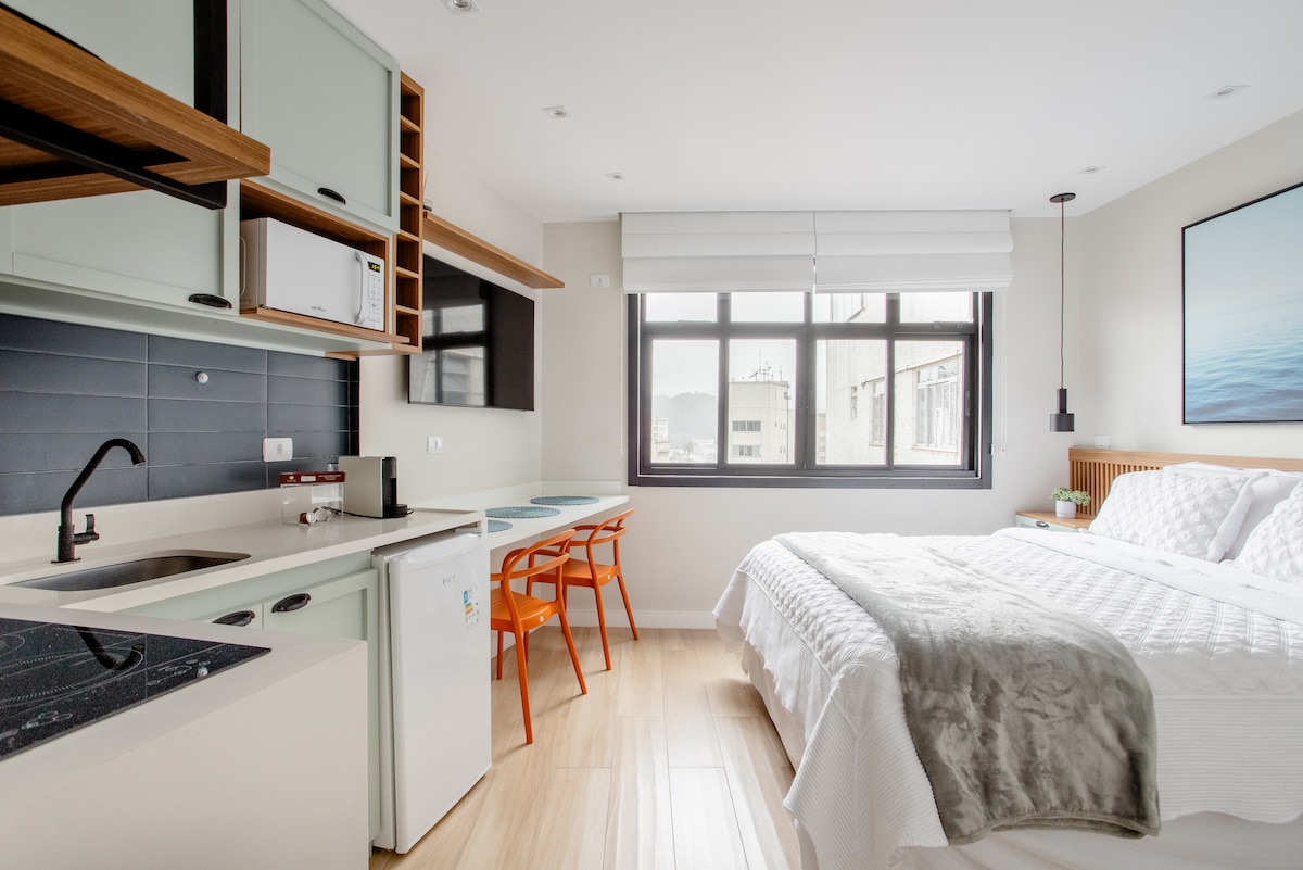 A modern studio showcases an integrated living space with a spacious bed, designed for comfort. A small kitchen area includes a refrigerator and microwave, while a dining table is complemented by bright orange chairs. Natural light illuminates the room through large windows.