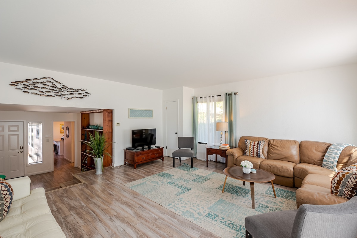 An open living room presents a comfortable seating area with a tan sectional sofa and a coffee table at its center. Natural light floods the space through partially draped windows, creating a welcoming atmosphere. A television is positioned on an entertainment unit, with decorative accents adorning the walls.