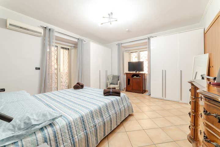 Пространа спална соба со телевизор, балкон и прозорец, многу светлина влегува во собата.