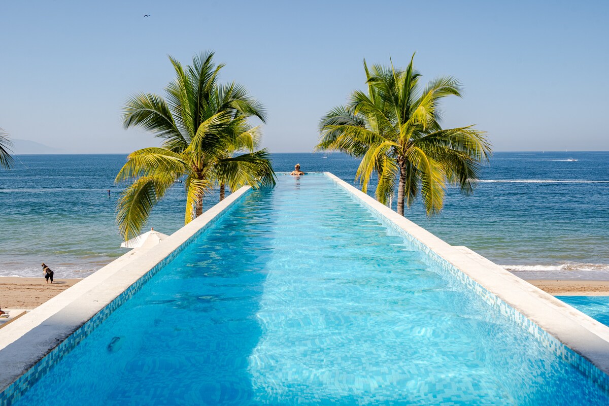 An infinity pool stretches toward the horizon, surrounded by palm trees. The clear blue water blends with the ocean, creating an uninterrupted view. The beach is visible in the background, with gentle waves lapping at the shore under a clear sky.