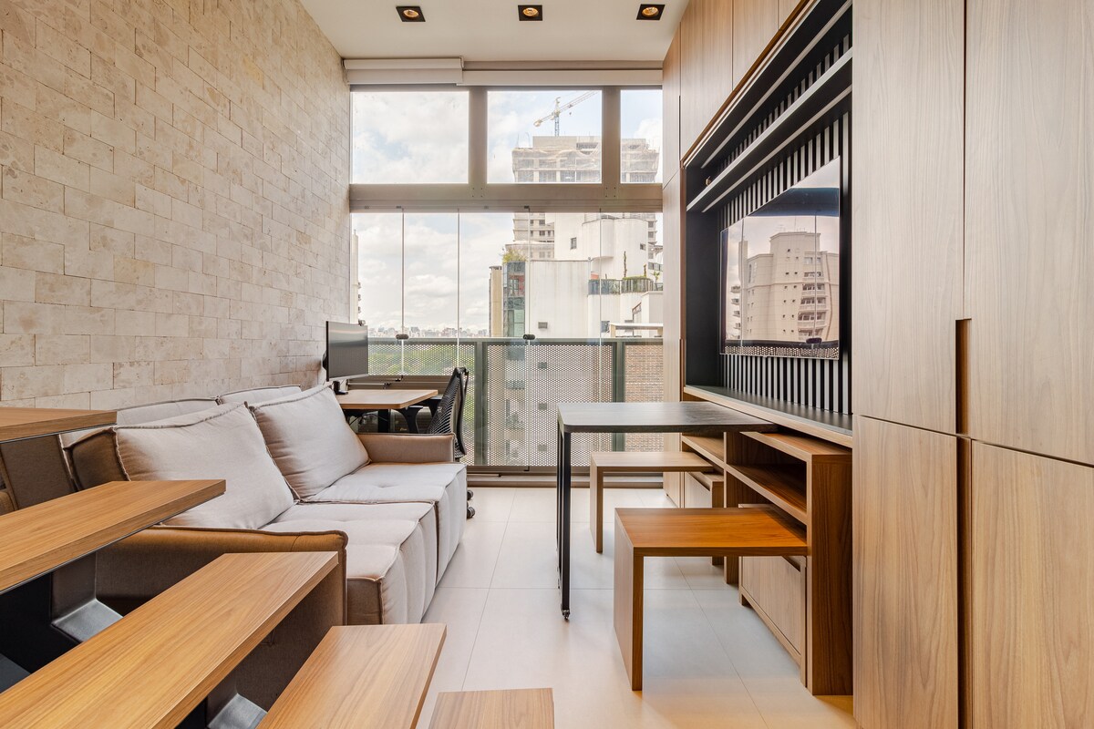 The living area is equipped with a sleek sofa and a wooden desk facing large windows that provide ample natural light. A monitor is positioned on the workspace, complementing a clean and modern aesthetic. Built-in cabinetry offers additional storage.
