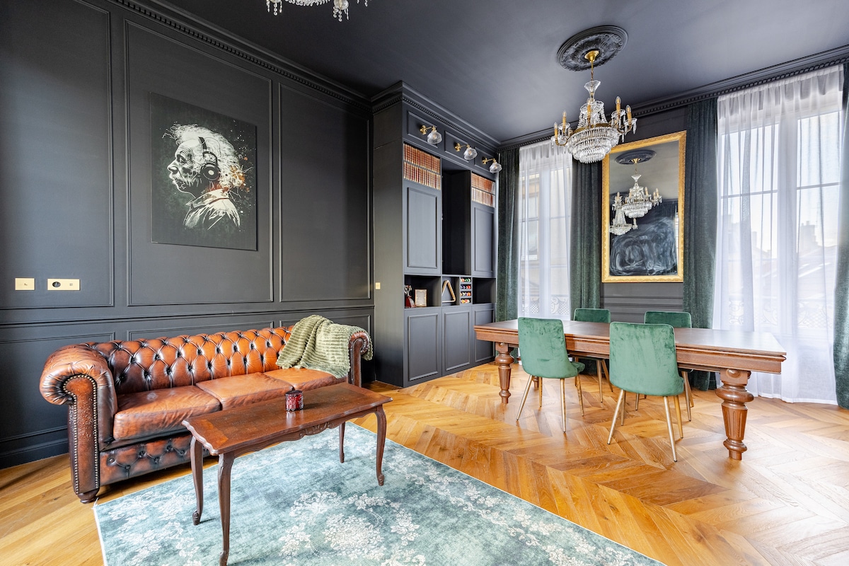 A deep brown Chesterfield sofa is positioned in the room, paired with a wooden coffee table. A sleek dining table is surrounded by green upholstered chairs. Elegant chandeliers hang from a dark ceiling, and large windows allow natural light to brighten the richly decorated walls.