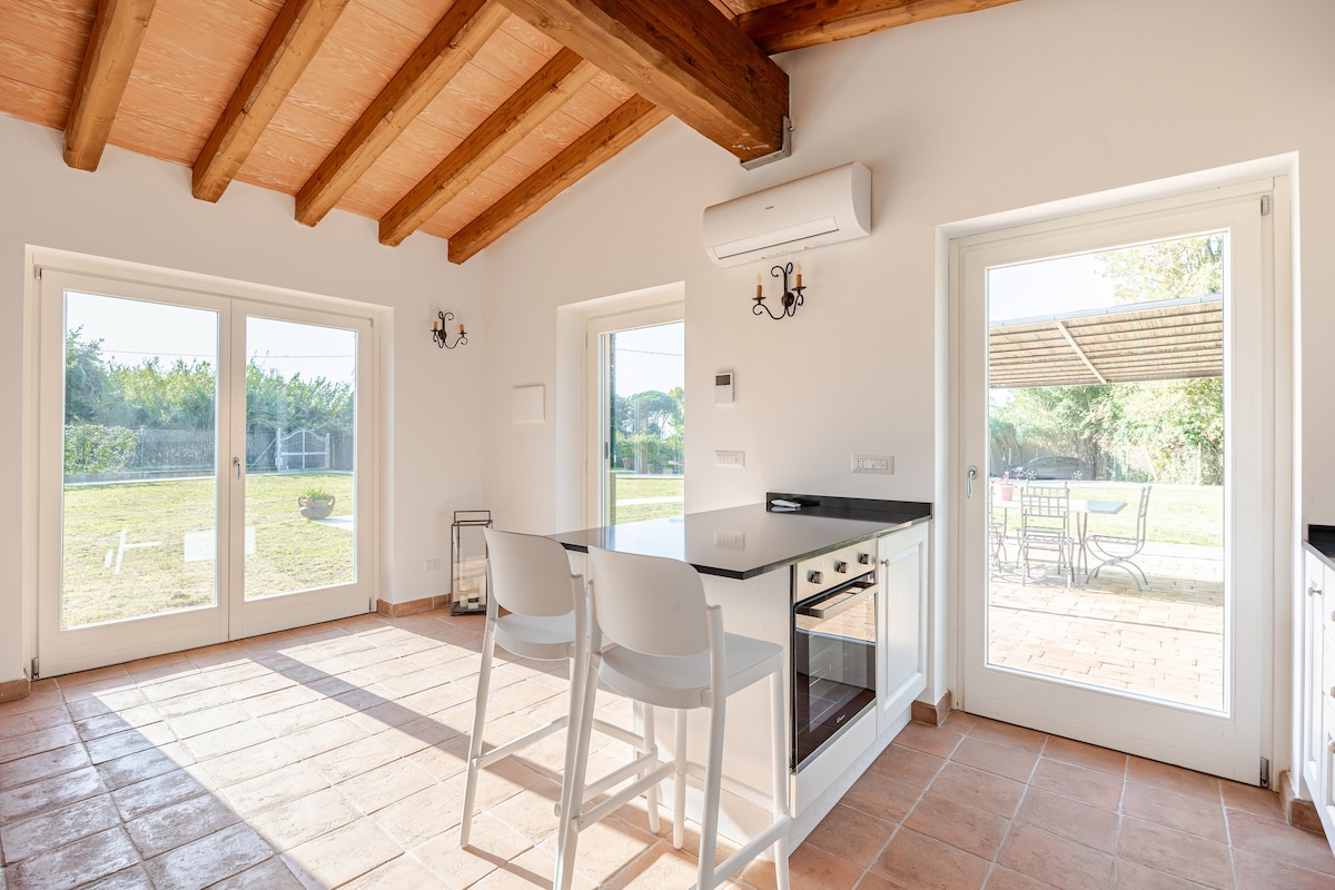 A well-lit kitchen area features a black countertop and a set of two white bar stools. Large glass doors open to a scenic garden view, allowing natural light to fill the space. Exposed wooden beams add character to the ceiling.
