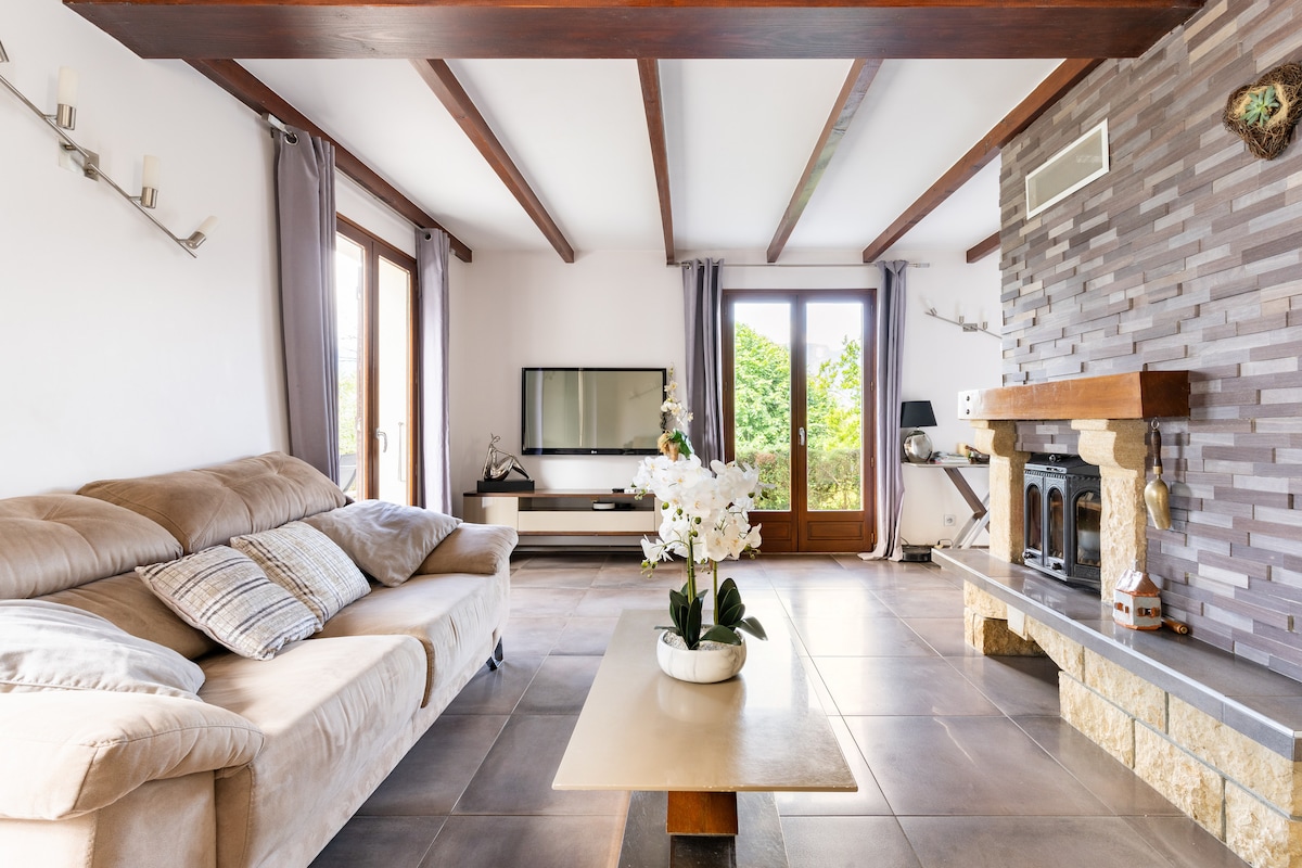 A spacious living area is presented with a comfortable sofa and a coffee table. Large windows allow natural light to fill the room, while a television and a decorative fireplace enhance the space. Soft curtains frame the windows, creating a serene ambiance.