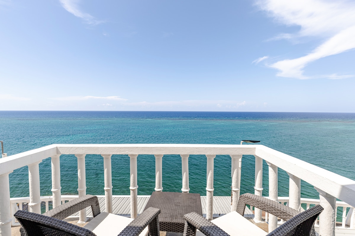 A private balcony features two comfortable chairs and a small table, offering unobstructed views of the expansive ocean. The bright blue water meets the clear sky, creating a serene backdrop ideal for enjoying morning coffee or sunset moments.