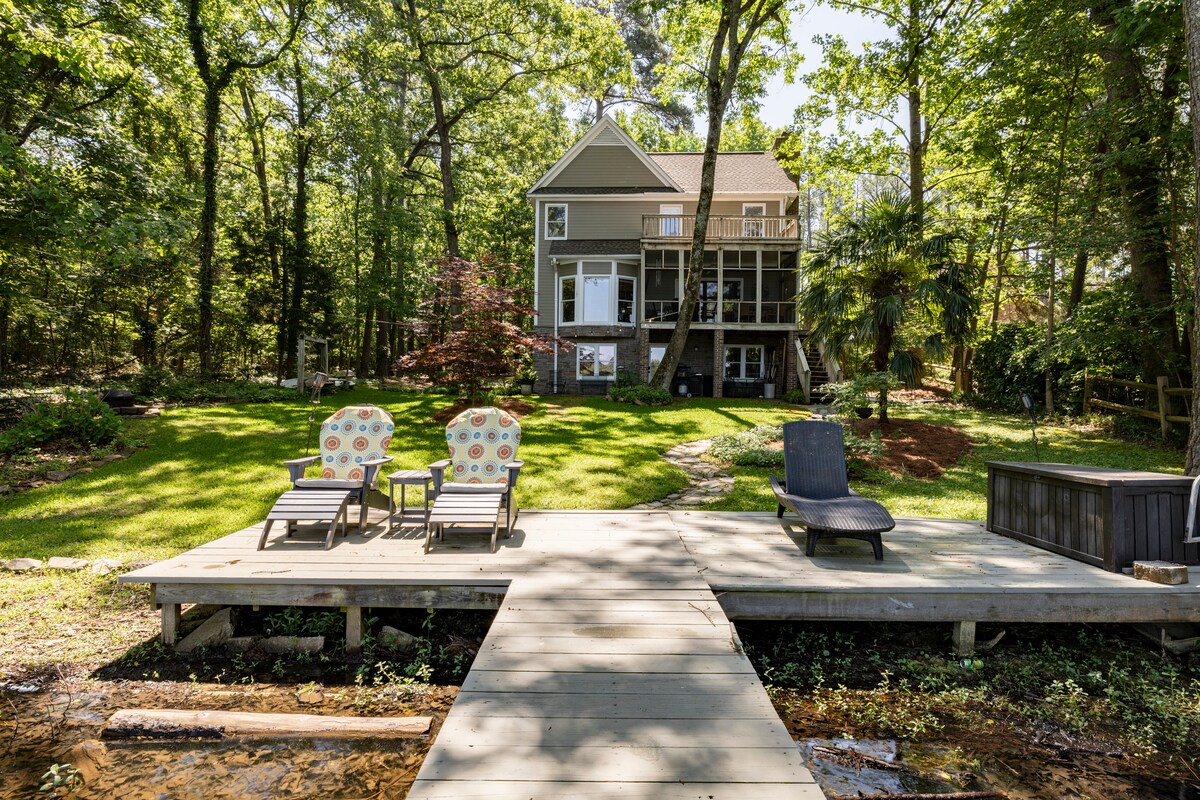 A wooden dock extends over the calm water, featuring two colorful chairs and a lounge chair. The house, surrounded by lush greenery, is partially shaded by trees, with a screened porch offering views of the lake. Soft sunlight filters through the leaves.