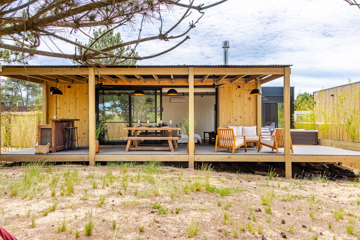The exterior of the home showcases a modern design in wood, featuring large glass windows. A spacious deck is equipped with a dining table and comfortable seating. Surrounding greenery and sandy areas contribute to a serene atmosphere.