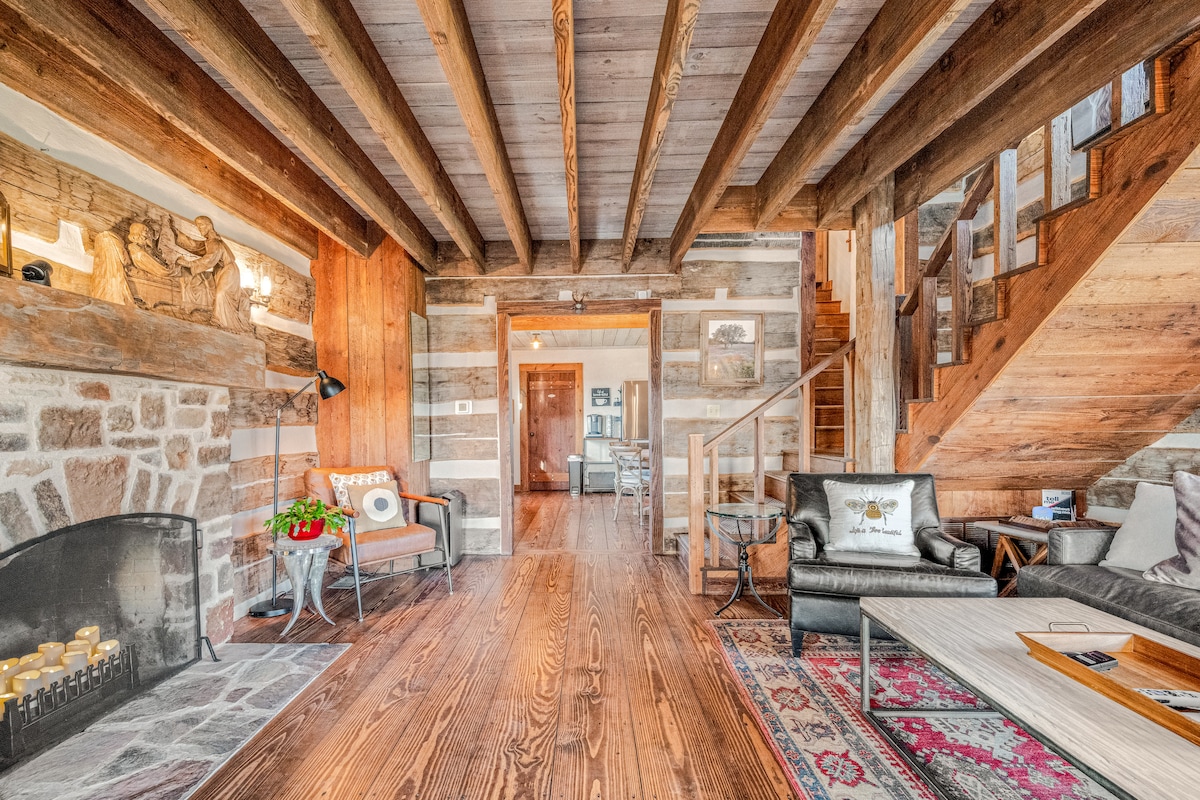The interior of the cabin features exposed wooden beams and rustic stone accents. A cozy seating area is arranged around a fireplace, complemented by a warm area rug. Stairs lead to the upper level, while a doorway opens to the kitchen and dining space.